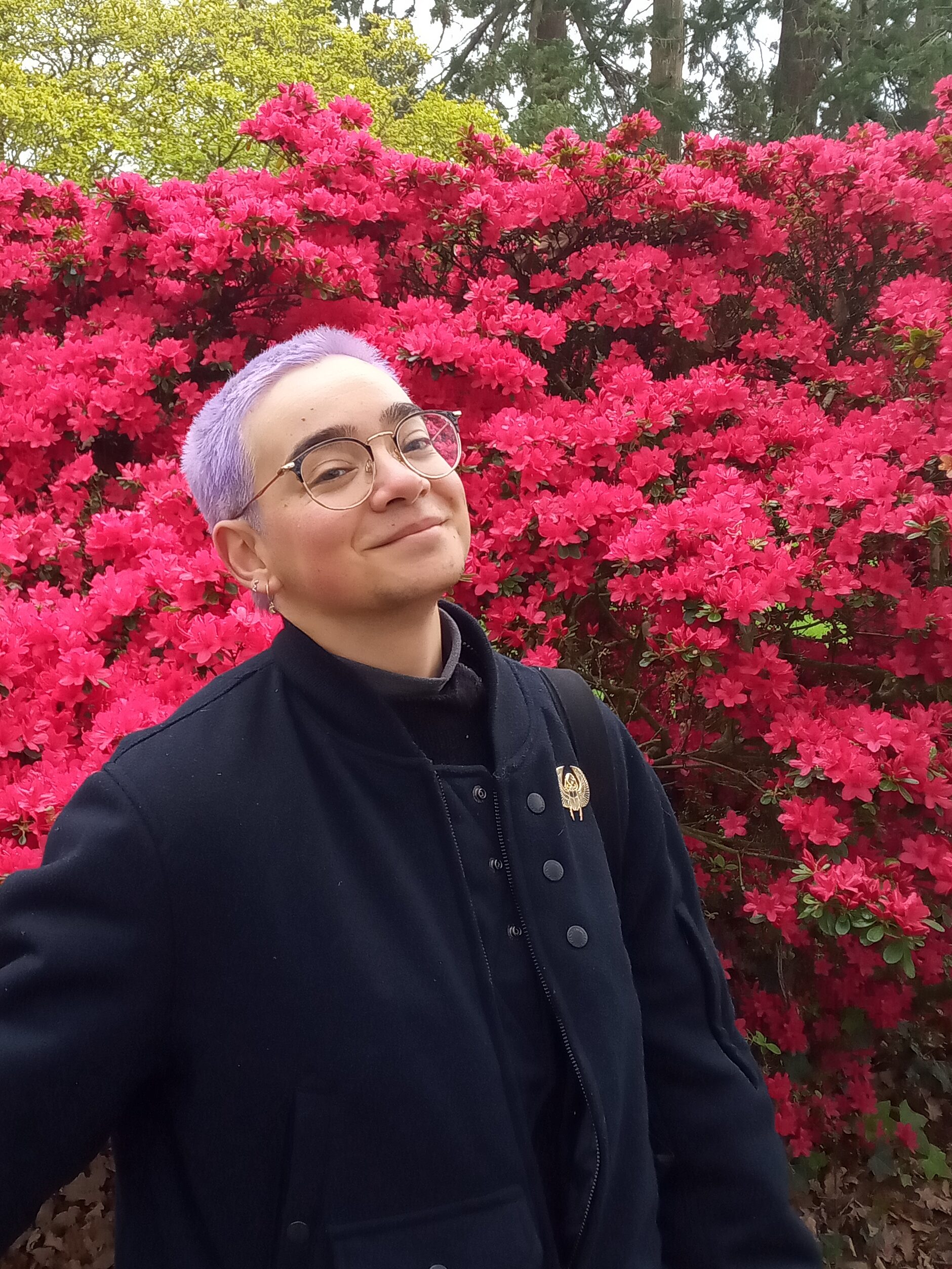 Personne en blouson, aux cheveux courts, devant un buisson de fleurs rouges.